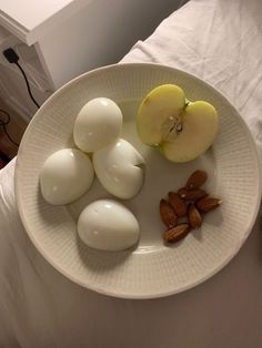 an apple, hard boiled eggs and almonds on a white plate with a bed in the background