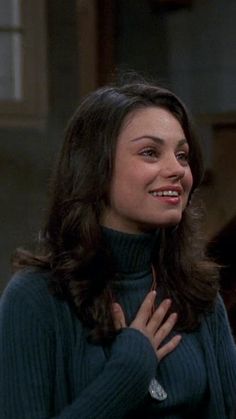 the young woman is smiling and holding her hands on her chest while wearing a turtle neck sweater