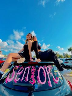 two women sitting on top of a car with the word senior spelled in pink letters