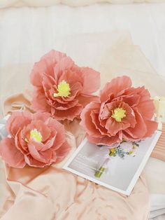 three large pink flowers sitting on top of a bed next to an open book and perfume bottle