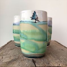 three ceramic cups sitting on top of a piece of wood with trees painted on them