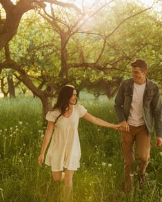 a man and woman holding hands walking through the grass with trees in the back ground