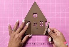 two hands are making a house out of cardboard and some scissors on a pink surface