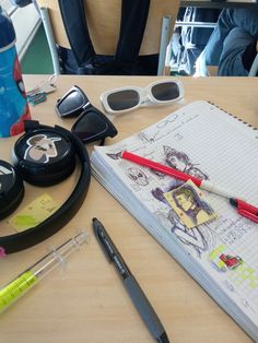 an open notebook sitting on top of a wooden table next to some pens and sunglasses