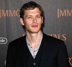 a man standing in front of a red carpet wearing a black suit and grey shirt