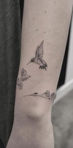 a small bird flying through the air with flowers on it's side forearm tattoo