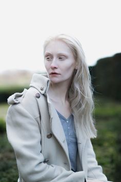 a woman sitting on top of a wooden bench wearing a coat over her shoulders and looking off into the distance