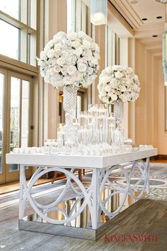 two tall vases with white flowers on top of a table in a room filled with windows