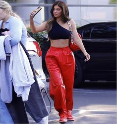 a woman in red pants and crop top walking down the street while looking at her cell phone