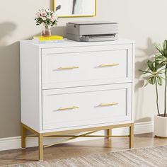 a white dresser with gold handles and drawers in a room next to a potted plant