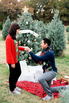 a man kneeling down next to a woman in front of a christmas tree on a blanket