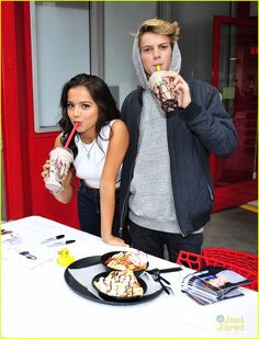 two people standing next to each other at a table with food and drinks in front of them