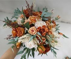 a bridal bouquet with orange flowers and greenery on the top is being held by someone's hand