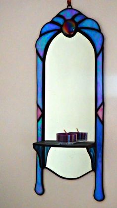 a stained glass mirror hanging on the wall next to a shelf with two candles in it