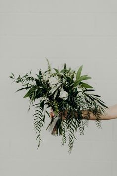 a woman holding a bouquet of flowers and greenery in her hand, against a white brick wall