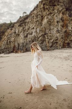 a woman in a long white dress walking on the beach next to a rock formation