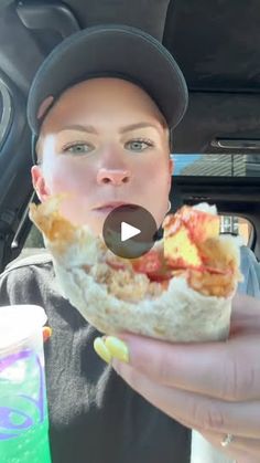 a woman holding up a half eaten sandwich in the back seat of a car while wearing a baseball cap