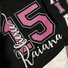 the back of a black and white shirt with pink letters on it, sitting on top of a carpeted floor