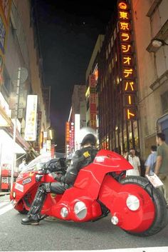 a man riding on the back of a red motorcycle down a street next to tall buildings