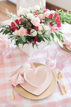 a table set with pink and white flowers in a heart - shaped vase, gold plateware, and napkins