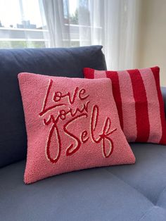 two pillows that say love your self on the back of a blue couch in front of a window