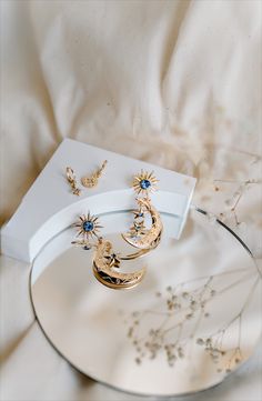 two gold earrings sitting on top of a glass plate next to a white box and flowers