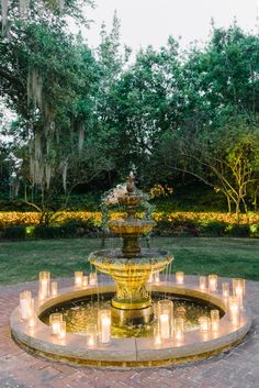 a fountain with lit candles in front of it and trees around the perimeter, surrounded by greenery