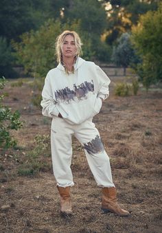 a woman standing in the middle of a field wearing white sweatpants and brown boots