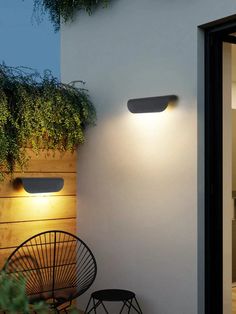 two lights on the side of a white wall next to a chair and planter