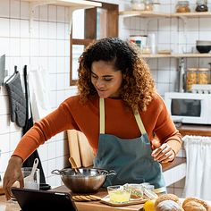 a woman in an orange shirt and blue apron is using a laptop on the kitchen counter