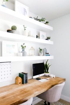 a wooden desk topped with a computer monitor next to a wall mounted bookshelf