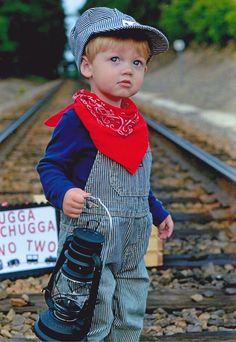 a young boy wearing overalls and a hat standing on train tracks with a sign in front of him