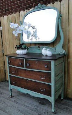 an old dresser with flowers and a mirror on it's top, sitting in front of a wooden fence