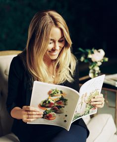 a woman sitting in a chair reading a cookbook while holding a plate of food