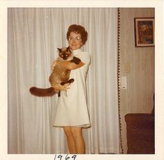 a woman holding a cat in her arms while standing next to a window with curtains