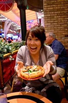 a woman sitting at a table with a pizza in front of her and pointing to the camera