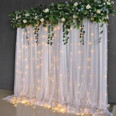 white flowers and greenery are hanging over the sheer curtain with fairy lights on it