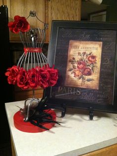 there are roses on the counter next to an old framed painting and wire vase with flowers in it