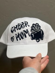 a baseball cap with the words cheer mom written on it in black and white, held up by a person's hand