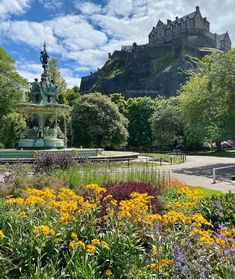 a fountain surrounded by flowers and trees in the middle of a park with a castle in the background