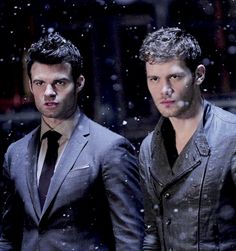 two men standing next to each other in front of snow falling on the ground and one man wearing a suit