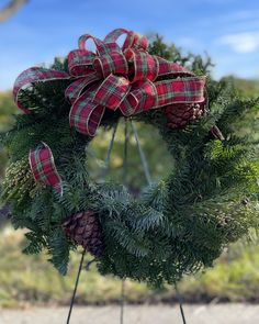 a christmas wreath with pine cones and plaid bow hanging on a tripod in the grass