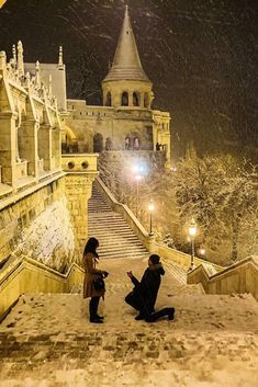 two people sitting on steps in front of a castle at night with snow falling all around