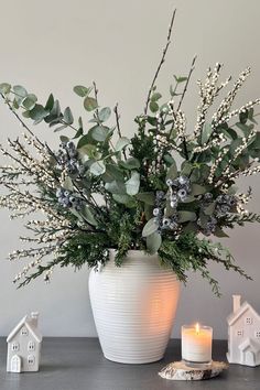 a white vase filled with lots of greenery next to a lit candle and small houses