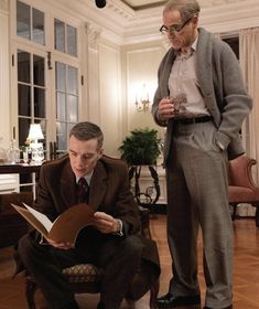 two men in suits look at a book while another man sits on a chair next to them