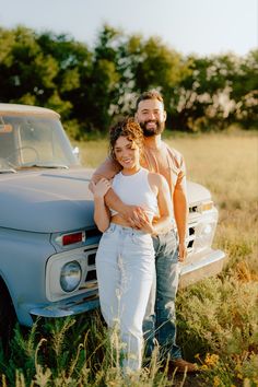 a man and woman standing next to an old pickup truck in a field with tall grass