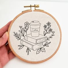 a hand holding a small embroidery hoop with coffee and books on it