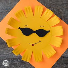 a paper plate sun with sunglasses on it sitting on top of a wooden table next to an orange piece of paper