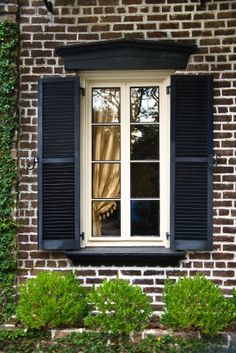 an open window with black shutters in front of a brick wall and shrubbery