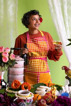 a woman in an apron holding a wine glass and standing next to a table full of food